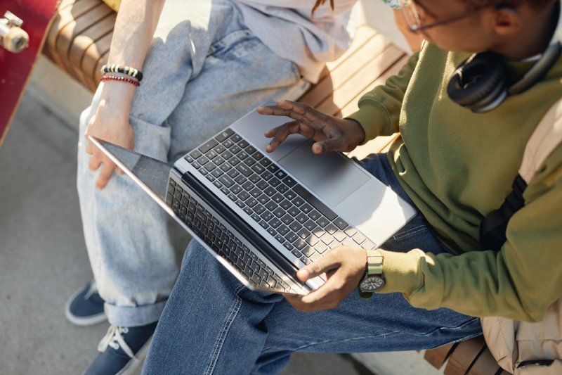 Black Teenage Boy Using Laptop Together with Friend on Bench Outdoors